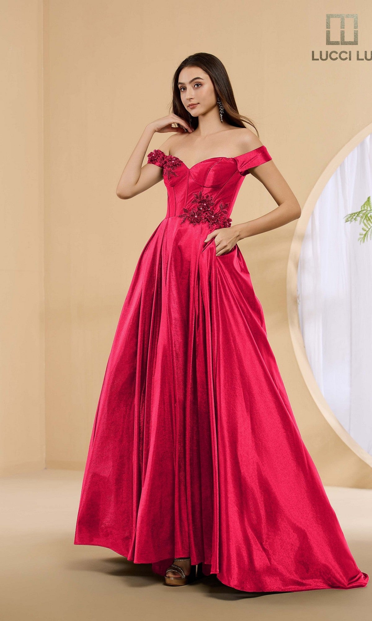 A woman stands in a neutral-toned room wearing the Lucci Lu 1472 long prom dress, an off-the-shoulder, floor-length A-line gown in wine with floral appliqué details.